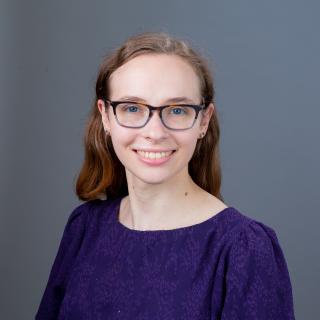 Headshot. Women with purple blouse and glasses on grey background. 