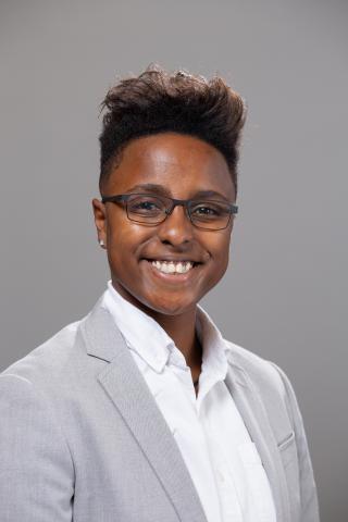 Jé is smiling at the camera in a grey blazer with a white button down shirt. They are wearing glasses with short, black hair.